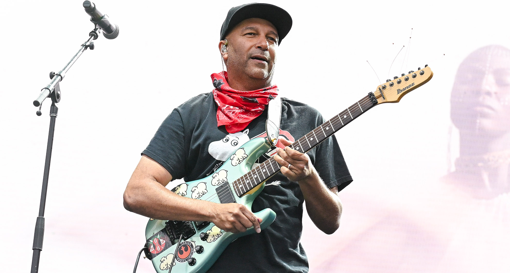 Tom Morello plays an Ibanez guitar against a bright backdrop. He wears a ballcap and a red bandana.
