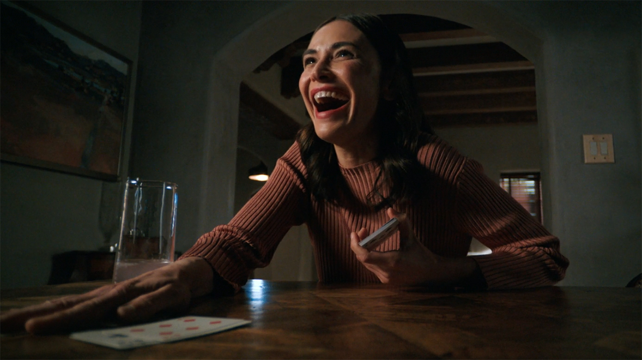 Zosia smiling wide and putting her right hand on a stack of table-based cards in Pluribus episode 8