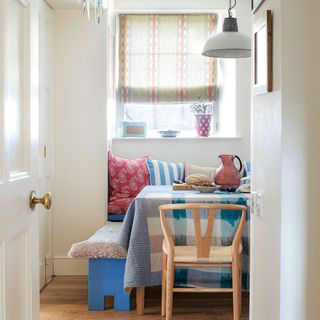 Corner of a seating area in a kitchen diner with checked tablecloth, striped window blind and mix of cushions and seating