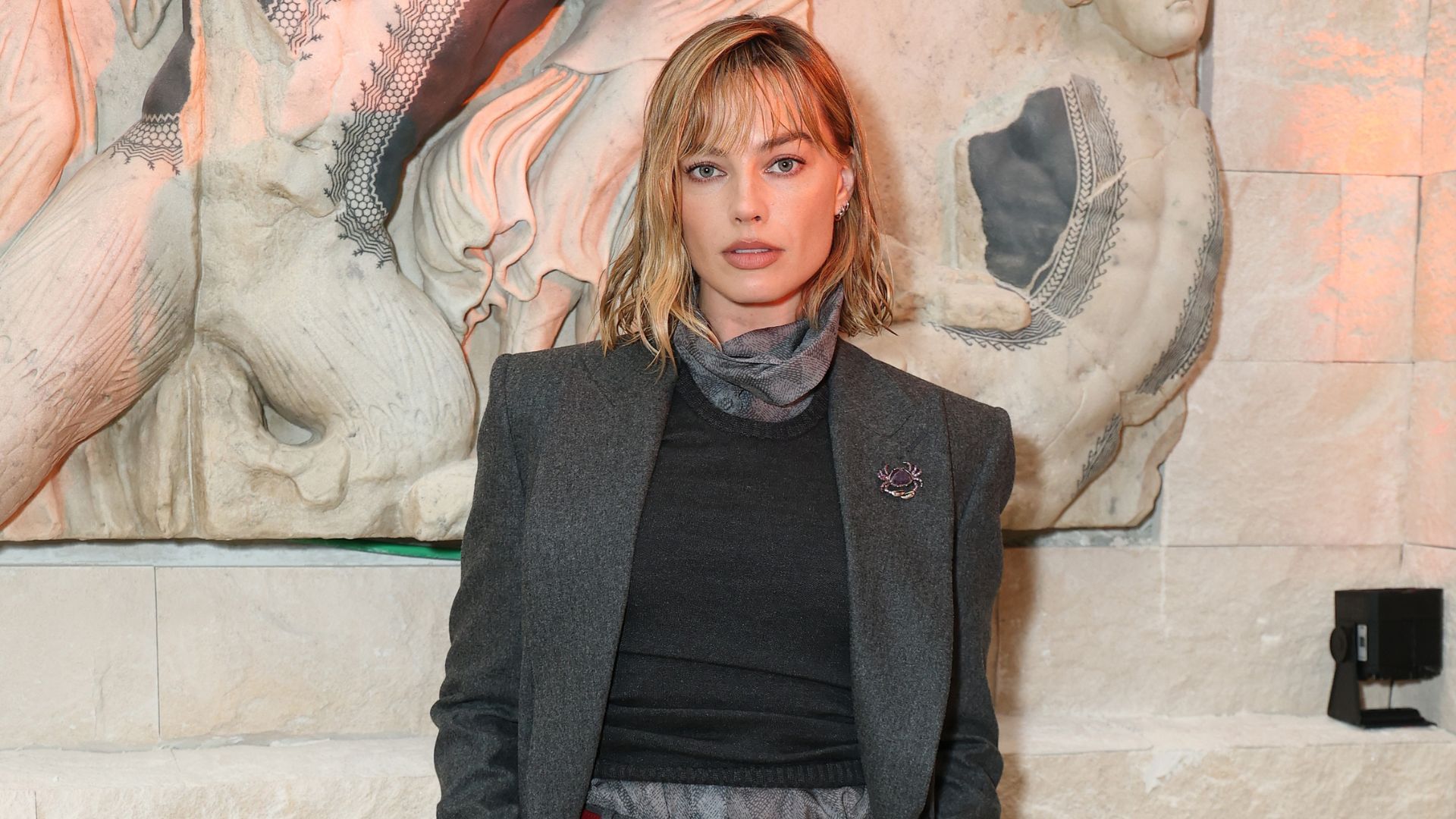 Margot Robbie wears burgundy flats with grey wool trousers and a grey longline blazer.