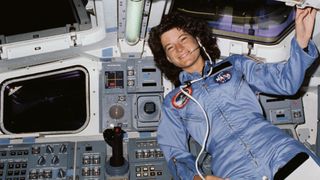 sally ride in a flight suit smiling and floating in the deck of the space shuttle with black space visible through the windows