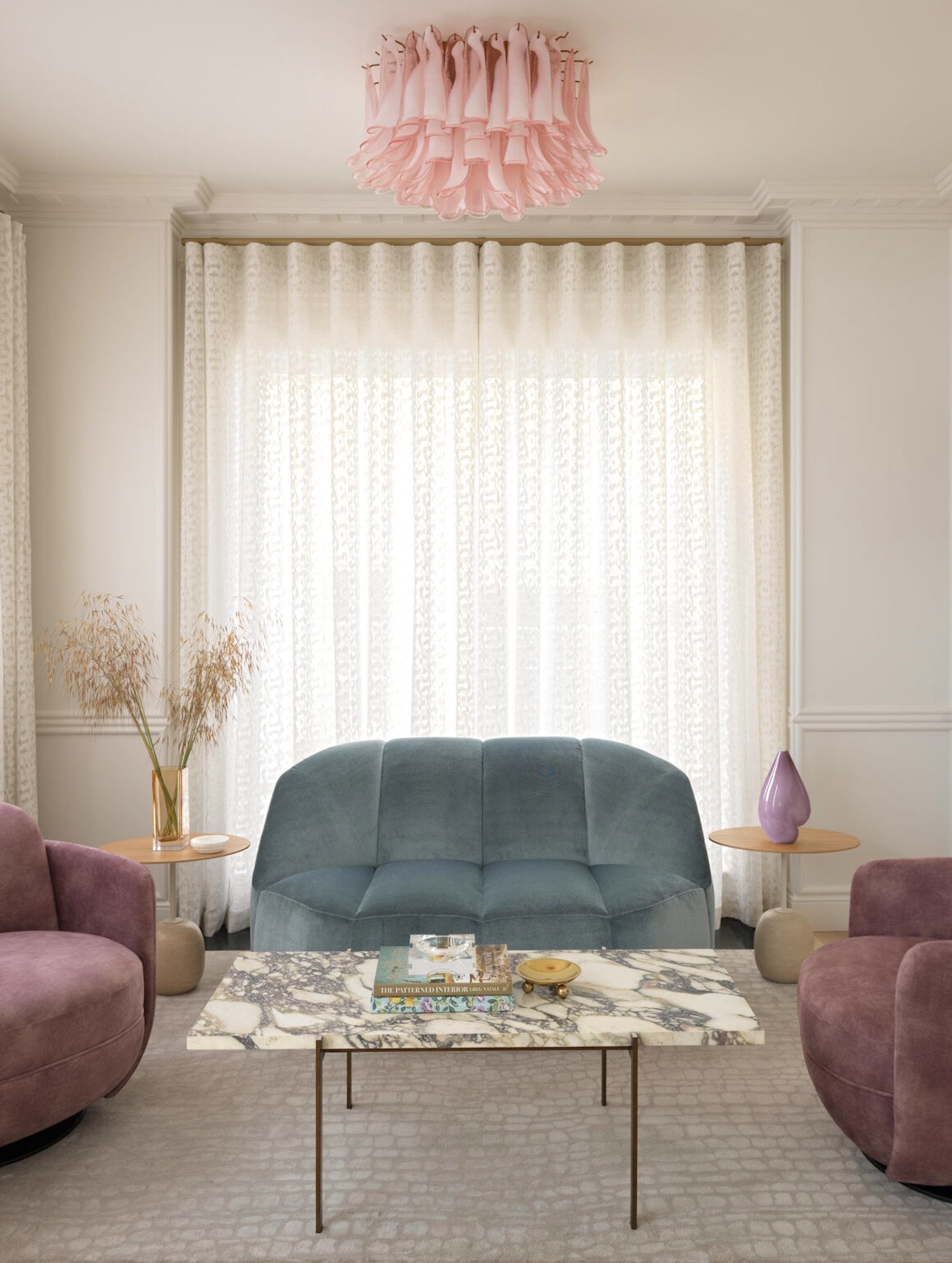 A soft neutral toned living space with a pink, modern style chandelier above the marble coffee table, and blue and pink toned sofas surrounding.