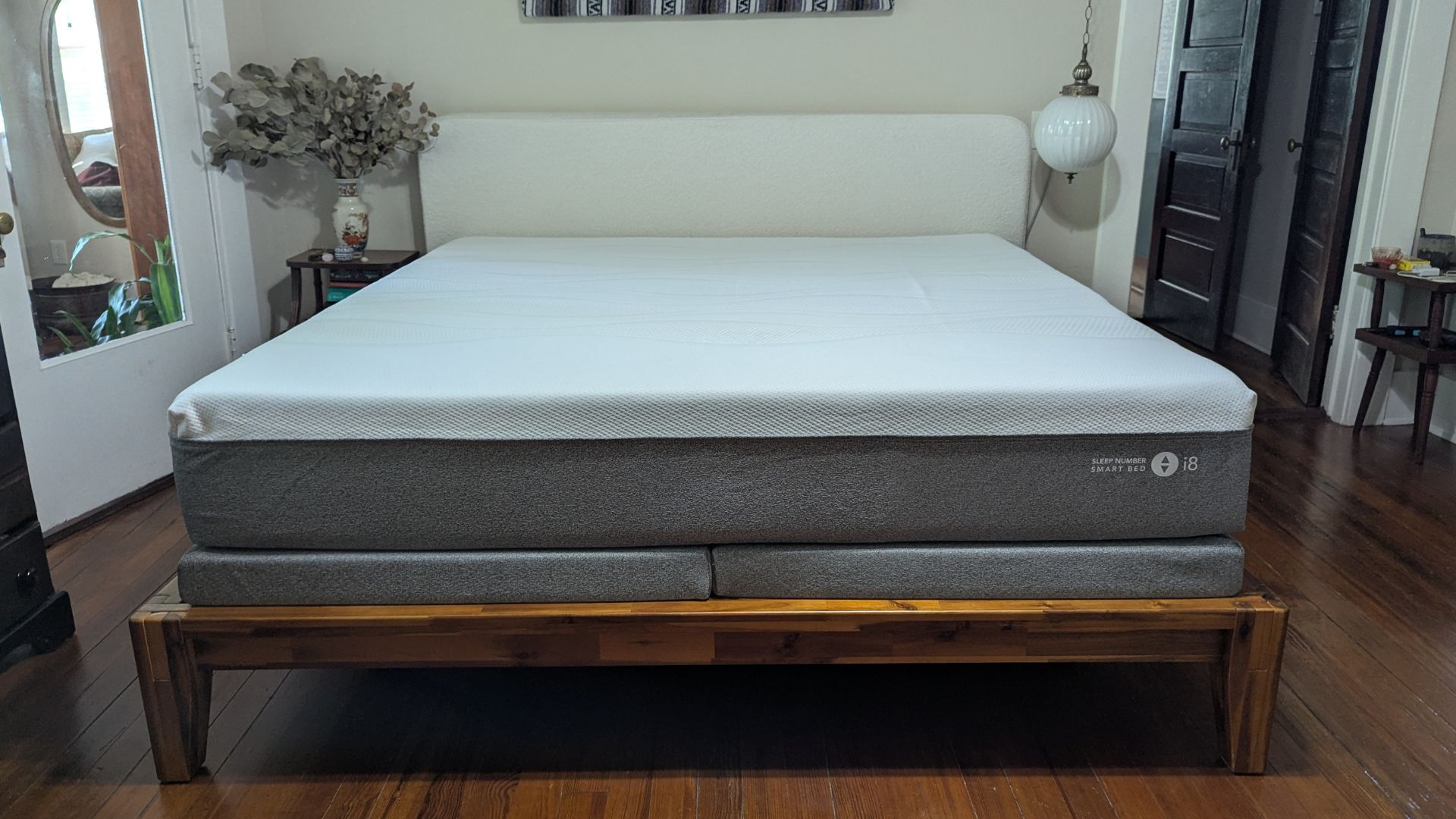 The image shows the Sleep Number i8 mattress on a wooden bed frame in a neutrally decorated bedroom