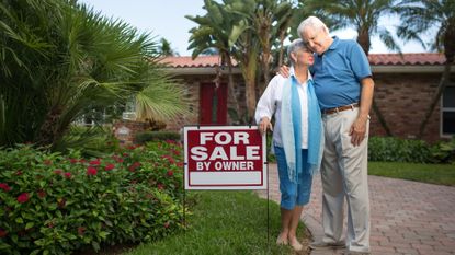 Sad senior couple standing in front of a house with a "For Sale" sign