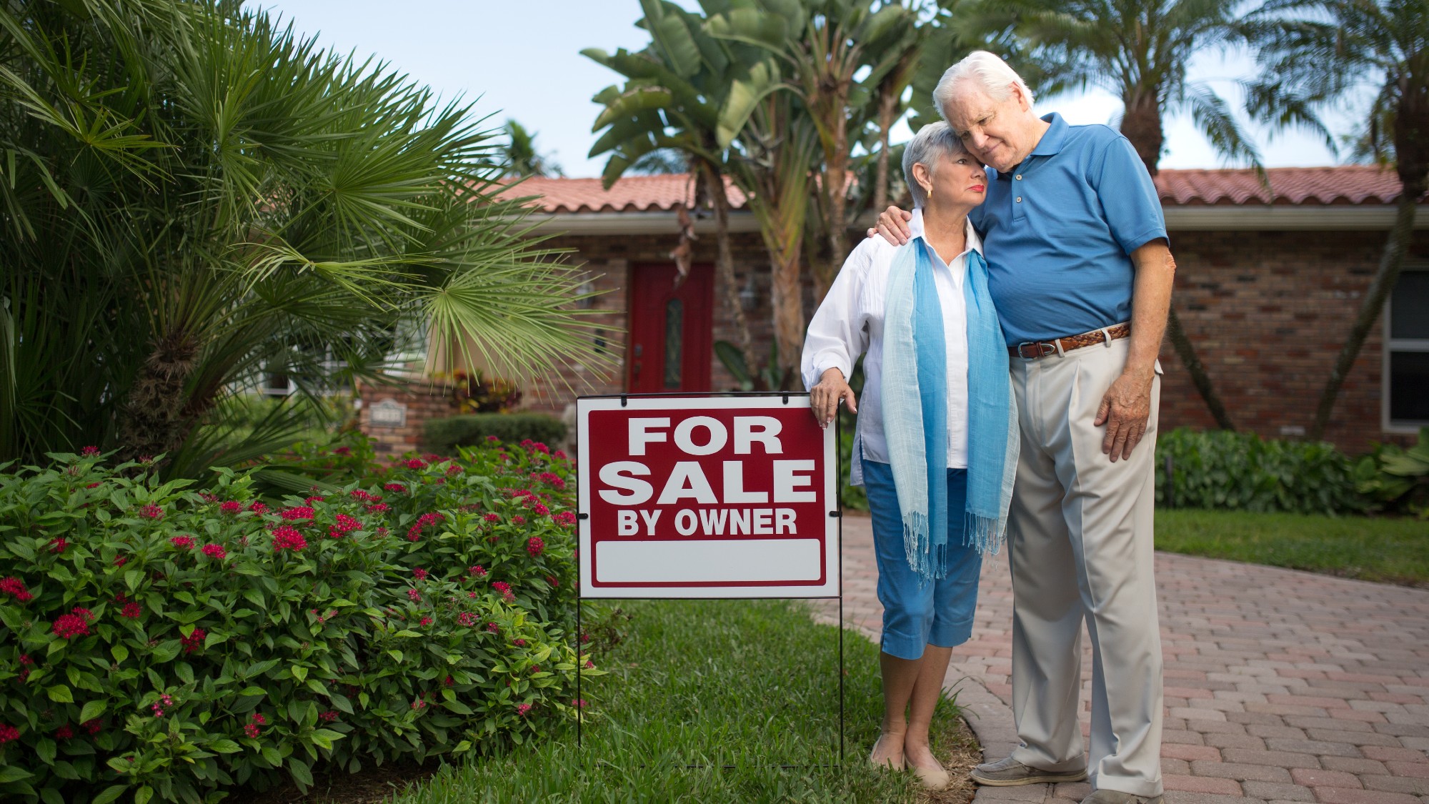 Sad senior couple standing in front of a house with a "For Sale" sign