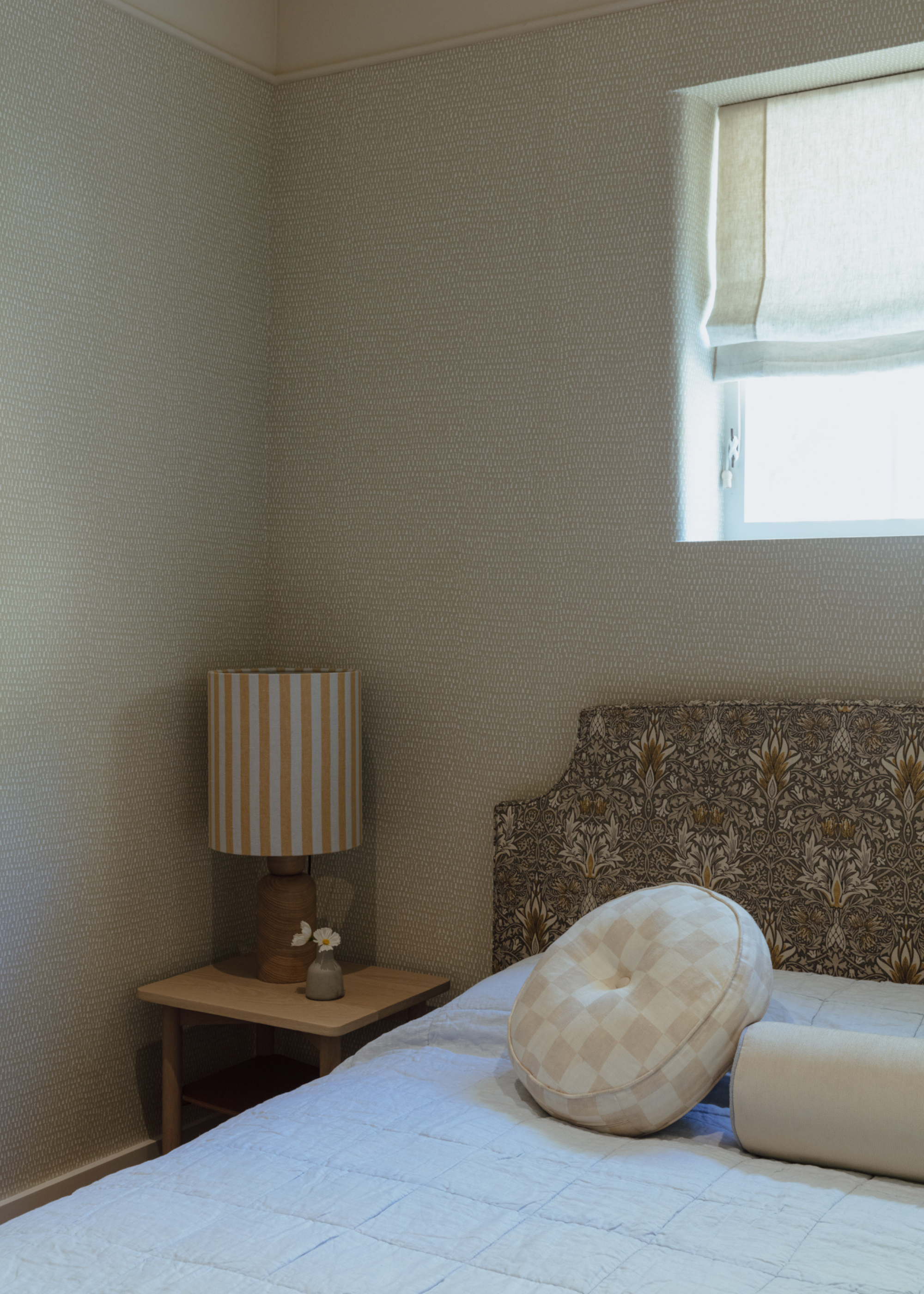 A bright bedroom corner with a wood side table and a striped lamp, a bed with a patterned headboard and a donut pillow, and a bolster pillow