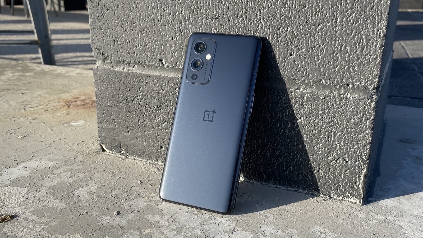 The back of a dark gray OnePlus 9, with the phone leaning against a wall.