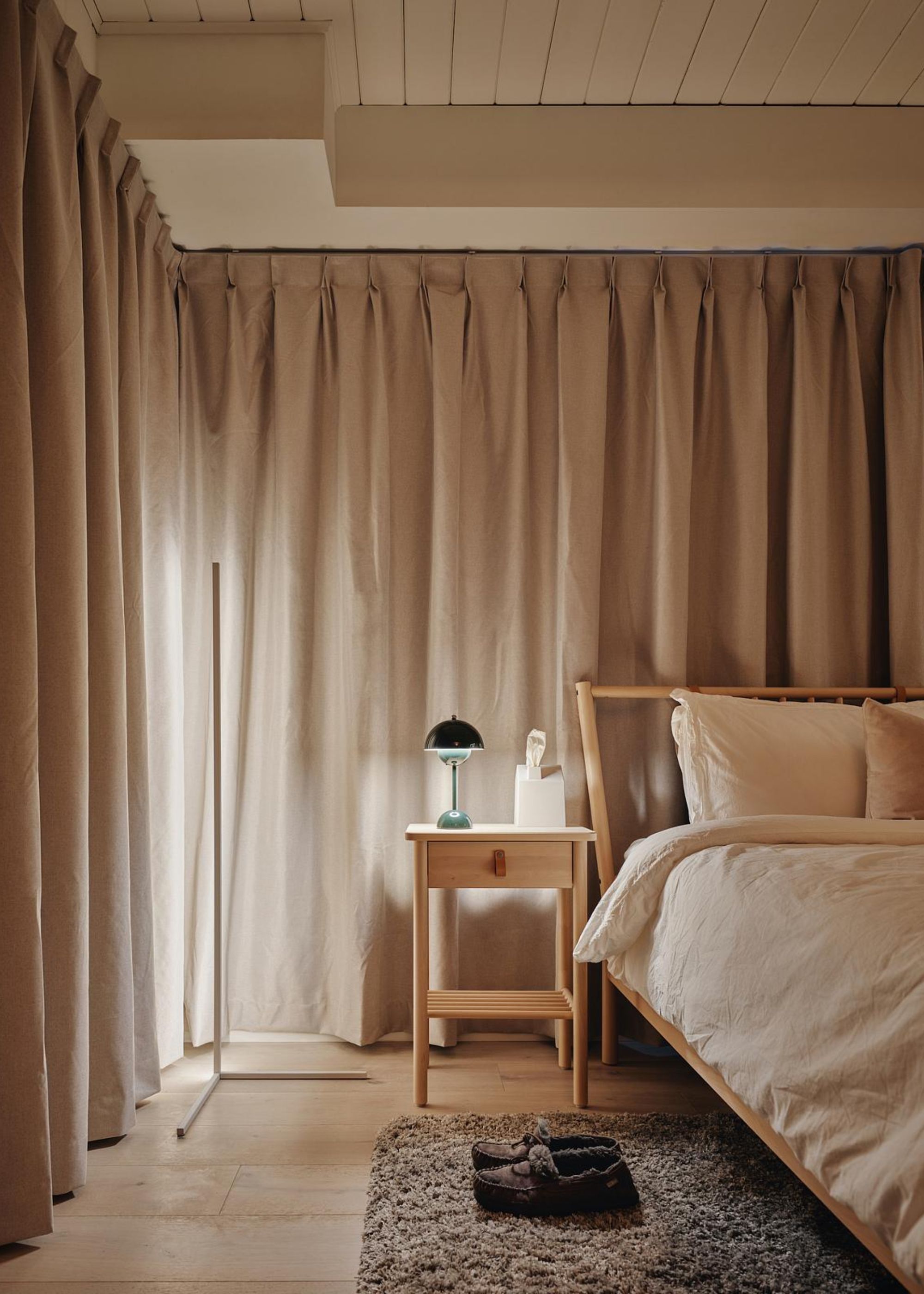 A beige and white bedroom with curtains draping all around. In the bedroom is a view of the bed with a side table beside it. On the side table is a table lamp. There are house slippers beside the bed.