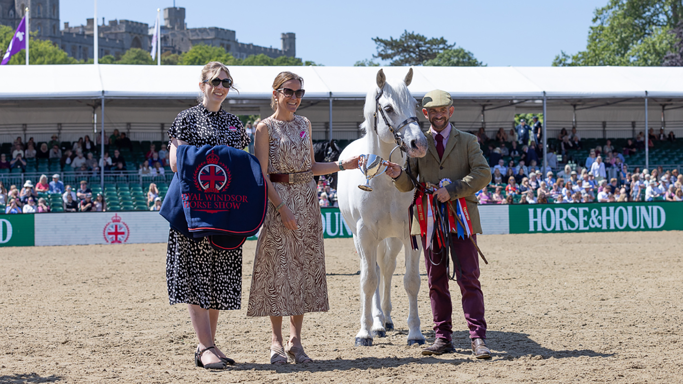 Horse &amp;amp; Hound&amp;rsquo;s Nicola McClure and Martha Terry present Connemara stallion Clover Hill Magic, handled by Jamie Frost, with a cup for winning the Horse &amp;amp; Hound Mountain and Moreland Supreme In Hand Championship at Royal Windsor Horse Show with the Windsor Castle behind the spectator stands.
