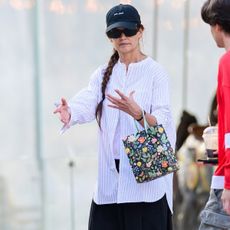 Katie Holmes wears a baseball cap, button-down shirt, black shorts, a floral bag, and brown leather loafers.