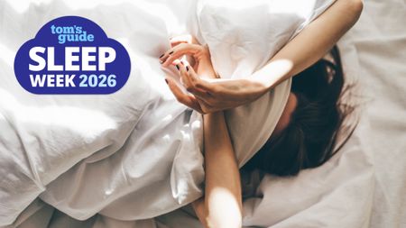 A woman with dark hair lies on her mattress with a white bed sheet covering some of her face because anxiety is keeping her awake and ruining her sleep. A blue Tom's Guide Sleep Awareness Week 2026 logo sits in the top left hand corner.