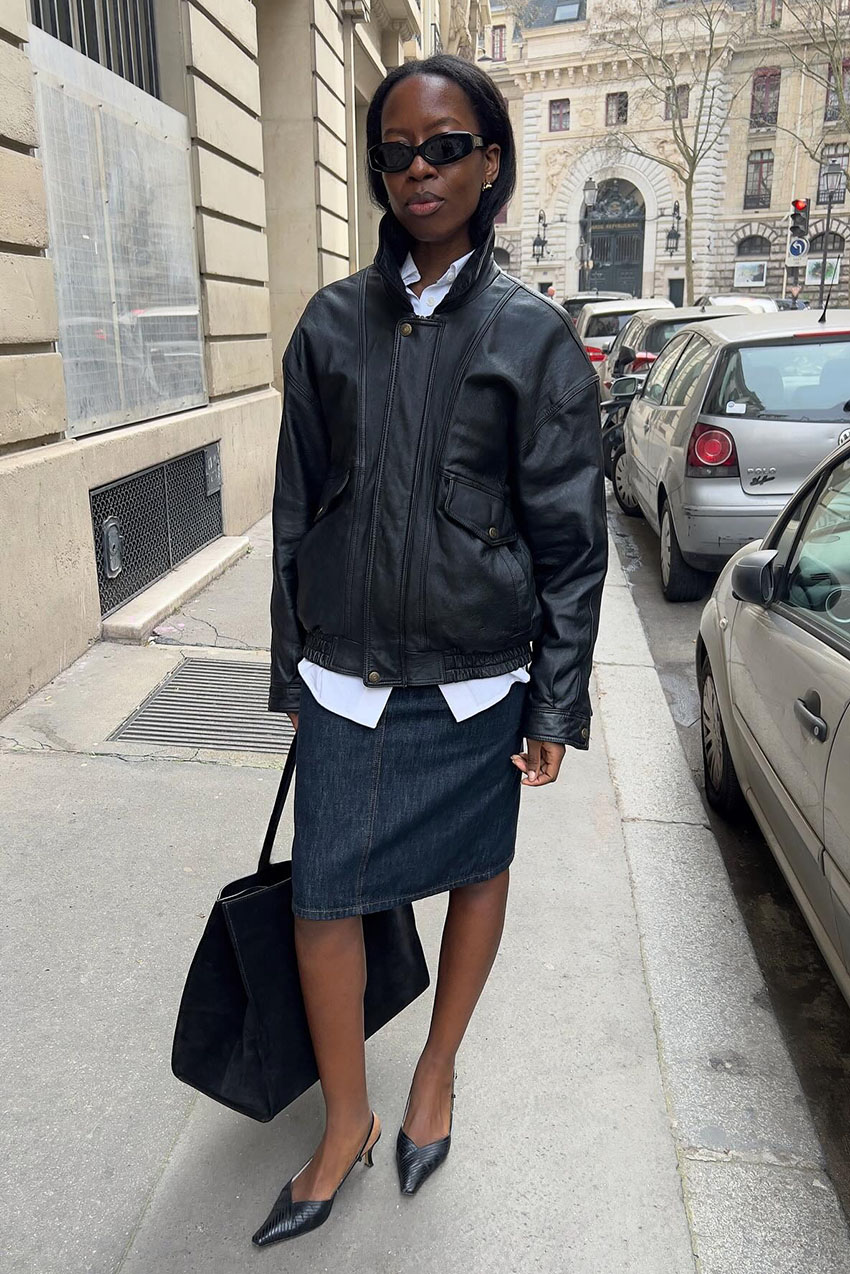 A woman wearing a leather black jacket, dark-wash denim skirt, and black slingback heels.