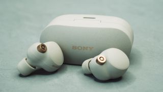 The Sony WF-1000XM4 earbuds in white next to their charging case on a pale surface.