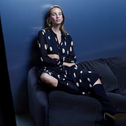 Model wearing a polkadot dress sold at Whistles with knee high boots sat on the back of a sofa