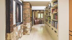 A library hallway space with bookshelves down one side, and an exposed brick wall on the other side with a Tudor-style window. At the end of the corridor is a pink upholstered chair under a window.