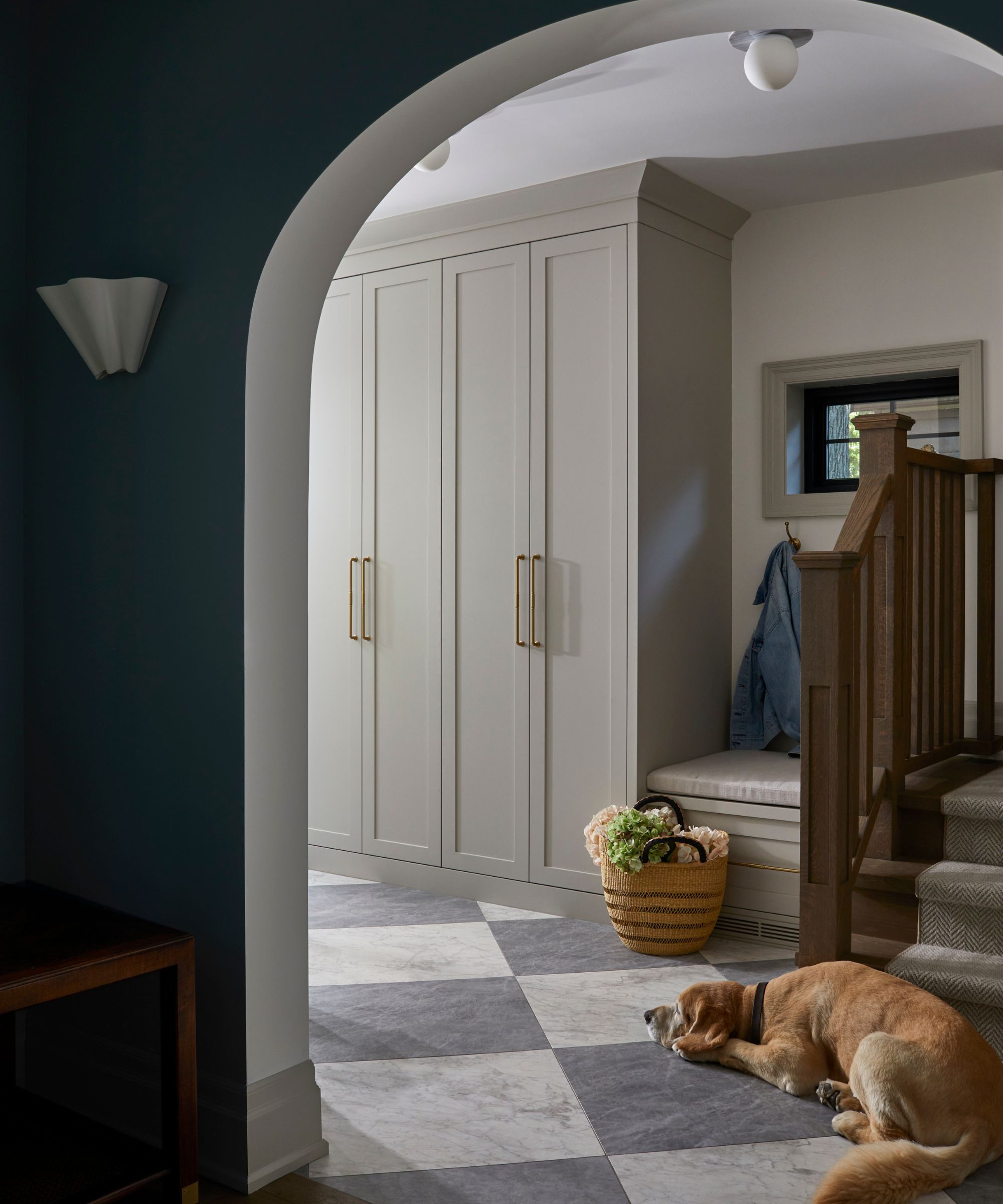 a large arched doorway into an entryway with checkerboard floor, custom joinery and a dog lying on the floor by a wooden staircase