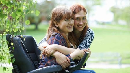 A woman in a wheelchair hugs her friend as they both smile in an urban park.