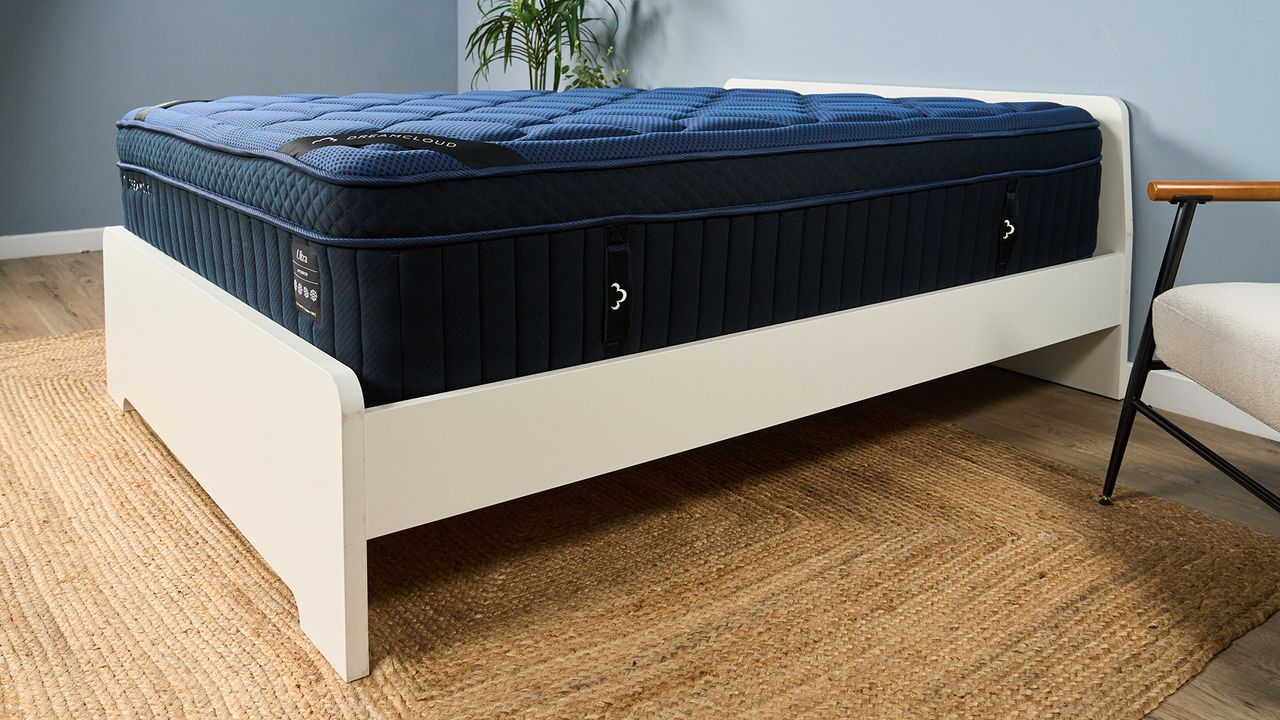 The image shows the close-up details of the DreamCloud Ultra Hybrid, a navy blue mattress placed on a white bed frame in a neautrally decorated bedroom