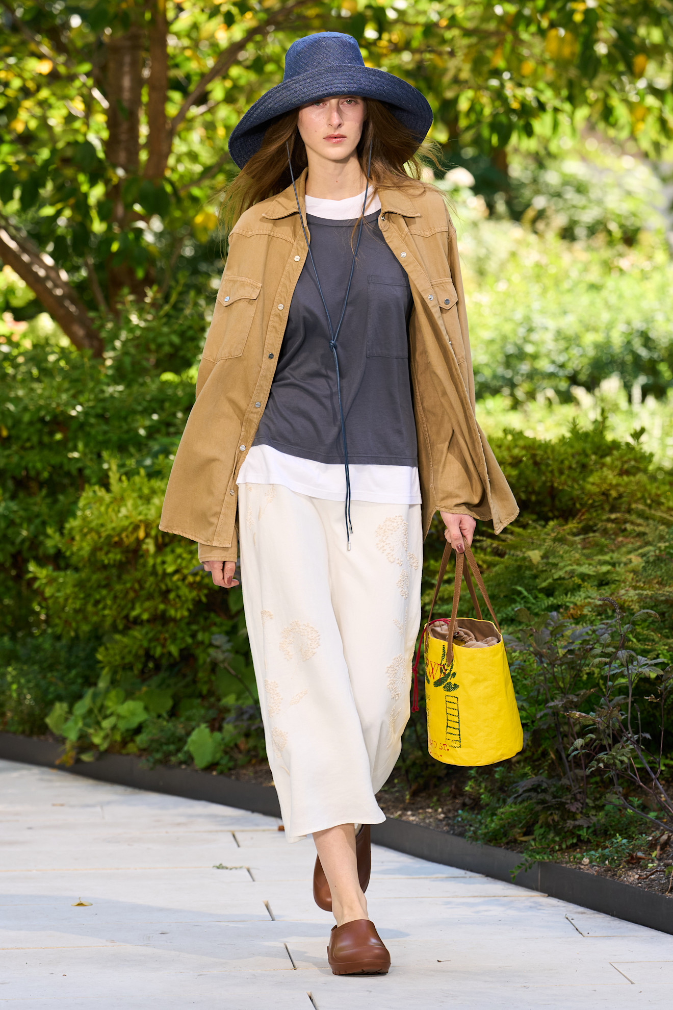 Layering staples for spring 2026 are shown in a photo from TWP's runway show, with a model walking while wearing a cargo button-down shirt styled on top of a white t-shirt layered under a gray t-shirt paired with white culottes, brown clogs, a blue bucket hat, and a yellow bucket hat