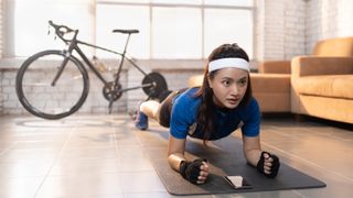 Woman wearing cycling kit performs the plank exercise in her living room. A bike connected to a turbo trainer is behind her and a smartphone is on the floor in front of her on the exercise mat 