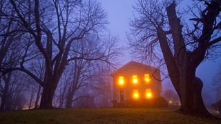 large house with all windows lit on large plot of land, misty fog and trees flanking either side of property