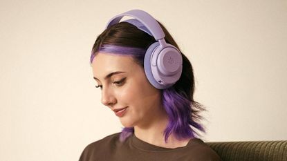 A young woman with a purple stripe in her dark hair wearing a lavender pair of SteelSeries Arctis Nova 3 Wireless Series headphones