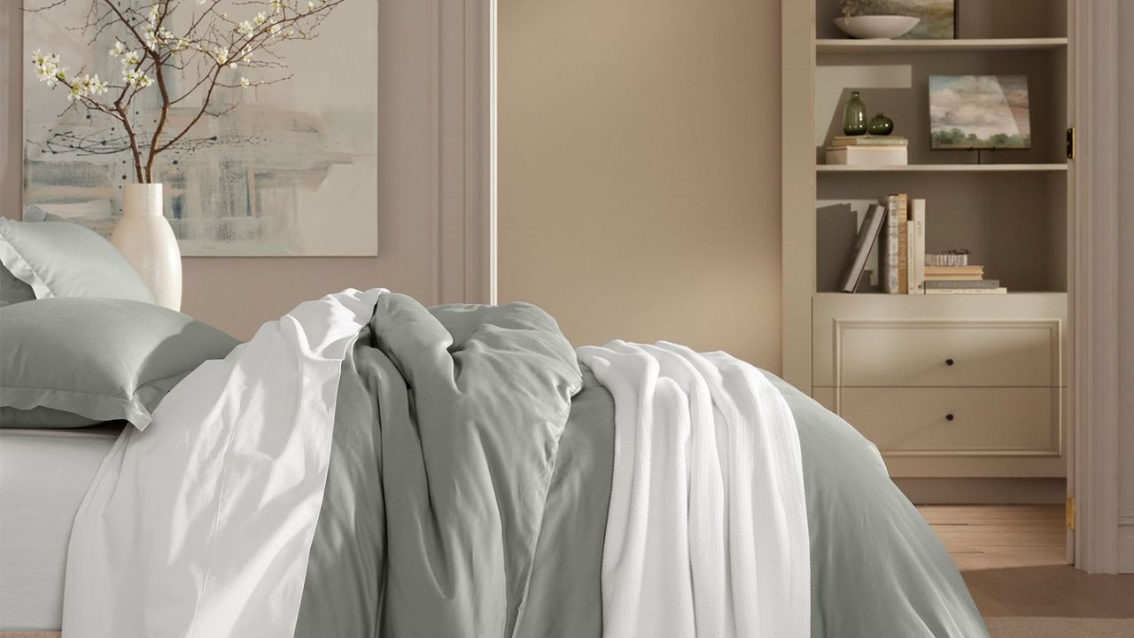 A gray comforter on a bed with white silk sheets against a beige wall.