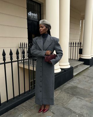 A woman wears a miu miu hat, gray long coat, alaia burgundy bag, and burgundy patent leather heels.