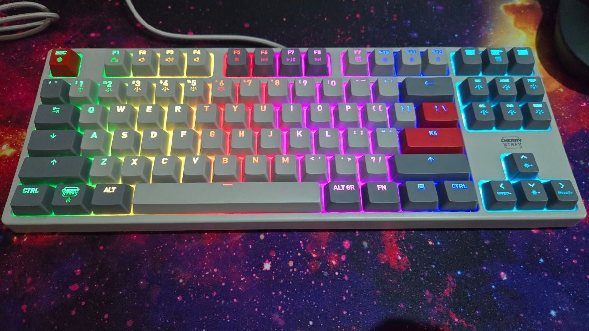 The Cherry Xtrfy K4V2 TKL gaming keyboard on top of a mouse pad depicting a nebula. The keyboard is grey with red accent keys, a grey braided wire, and the bright RGB lights switched on.