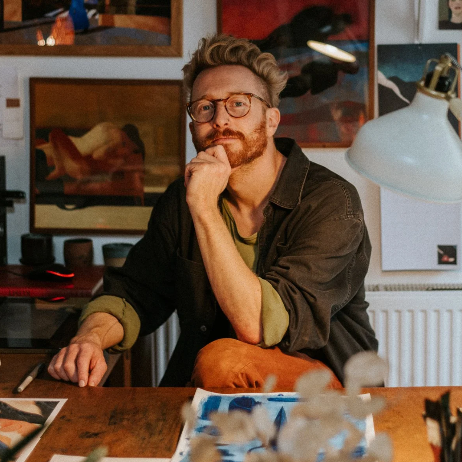 A man with short brown-blonde hair in a black shirt and a green t-shirt, perched in an art studio