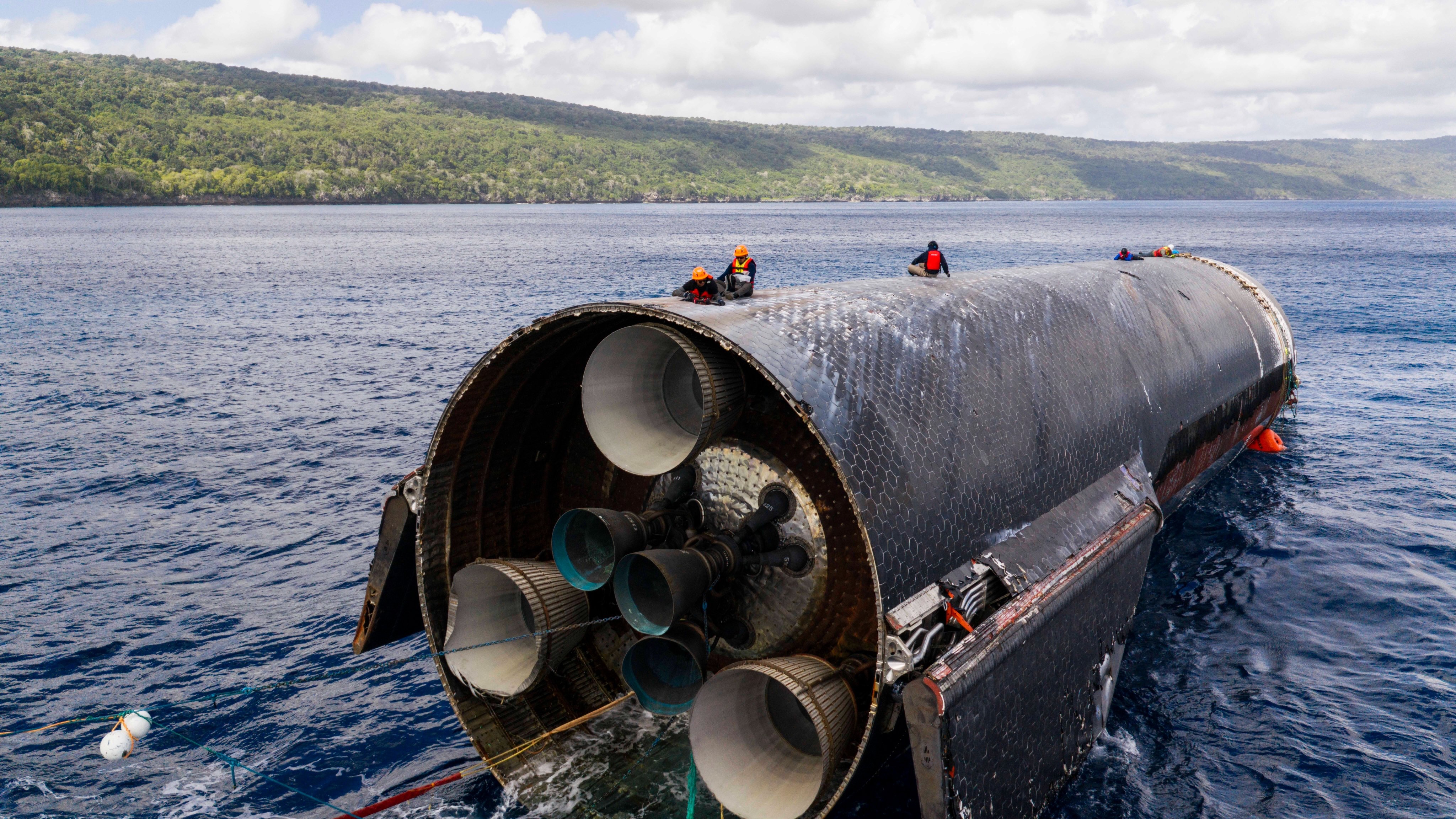 several people sit atop a large cylindrical black spacecraft floating in the ocean just off an island's coast
