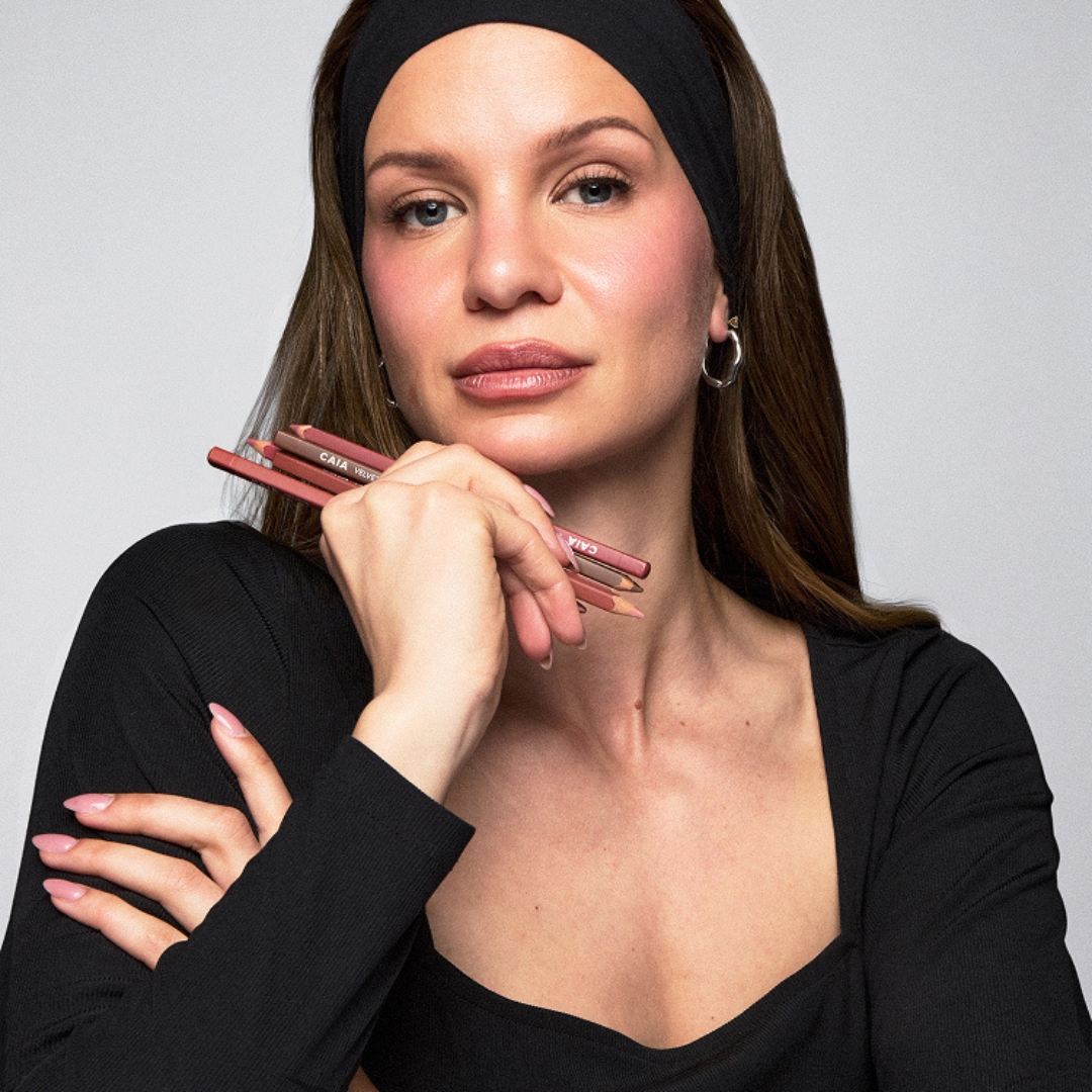 Image of Carolina Benita, Global Lead Makeup Artist at CAIA, with long brown hair, wearing a black headband and black square-neck top, holding CAIA lip liners