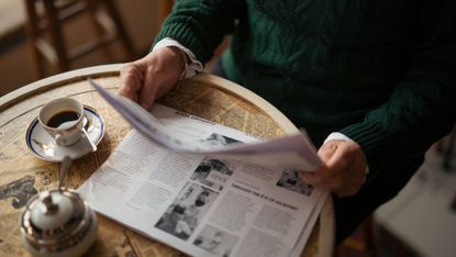 A view of a person in a green sweater holding up a newspaper on a table with a cup of coffee next to them