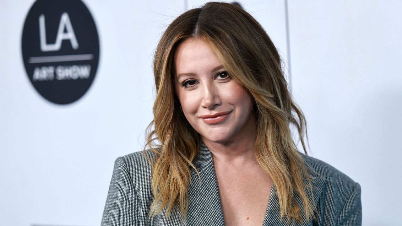 Ashley Tisdale, a white woman wearing a gray blazer, standing against a white background with a black circle