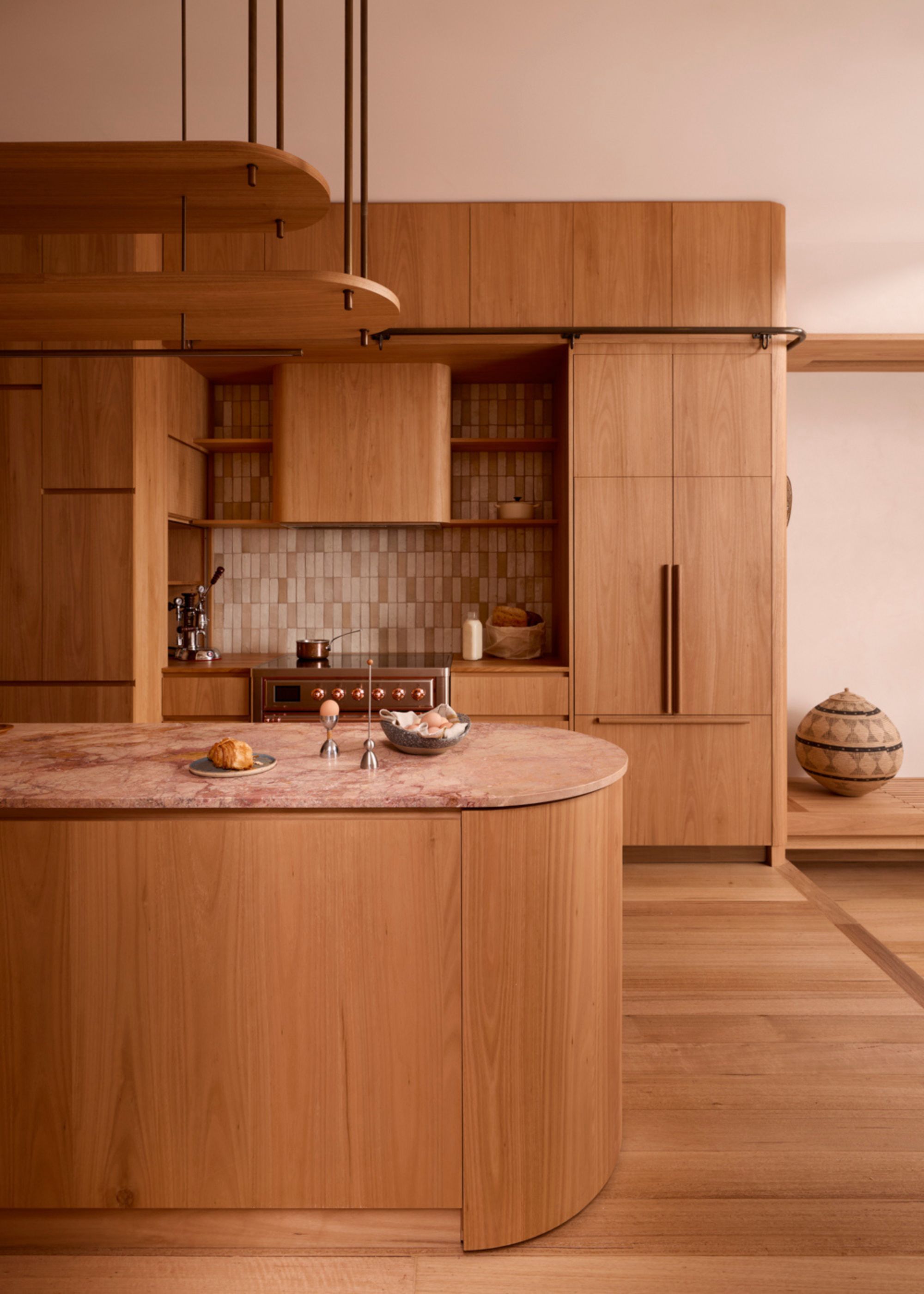 A kitchen with wood cabinets, a tiled backsplash, brass accents, an island with a stone top and a plate of pastries alongside a bowl of eggs