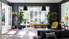 Grey living room, tall windows, window seat with yellow cushions, French floral fabric upholstered chair, oversized black ceiling lights, pale wood flooring, sofa, round antique table, house plants