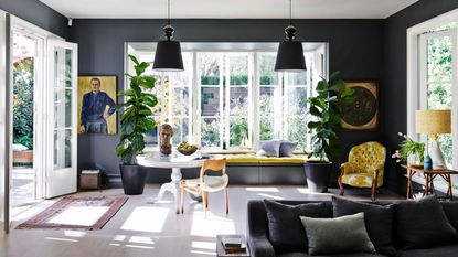 Grey living room, tall windows, window seat with yellow cushions, French floral fabric upholstered chair, oversized black ceiling lights, pale wood flooring, sofa, round antique table, house plants