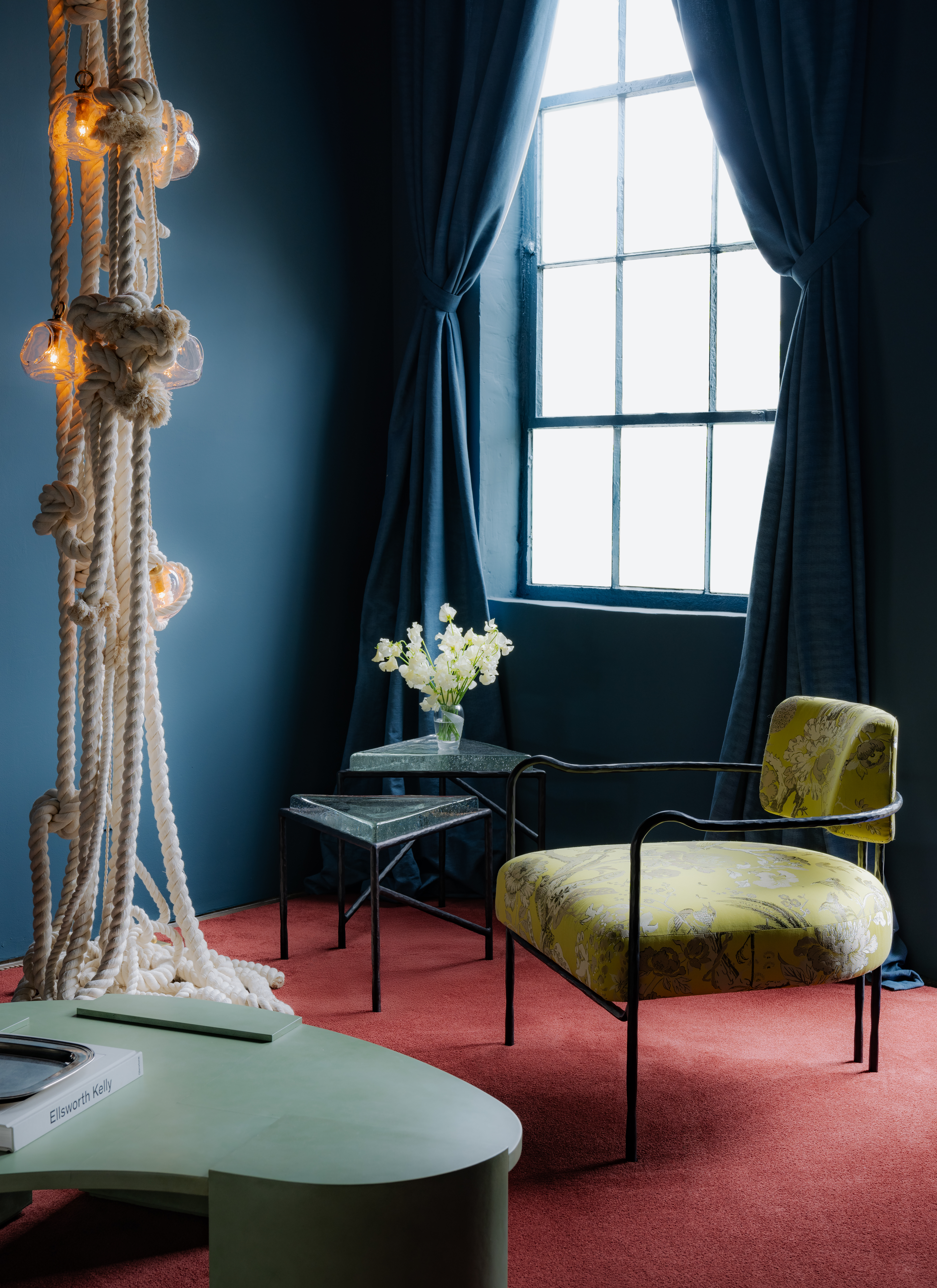 A warmly decorated, imaginative space features a red velvety carpet, a sculptural, pale green coffee table, an upholstered chair in wrought iron and yellow, floral fabric, blue walls and curtains, and a rope-made sculptural installation pending from the ceiling.