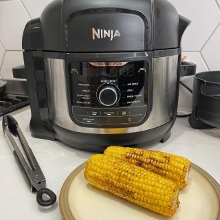 Ninja Foodi 9-in-1 Multi-Cooker behind cooked corn on the cob next to silver tongs on a white kitchen counter