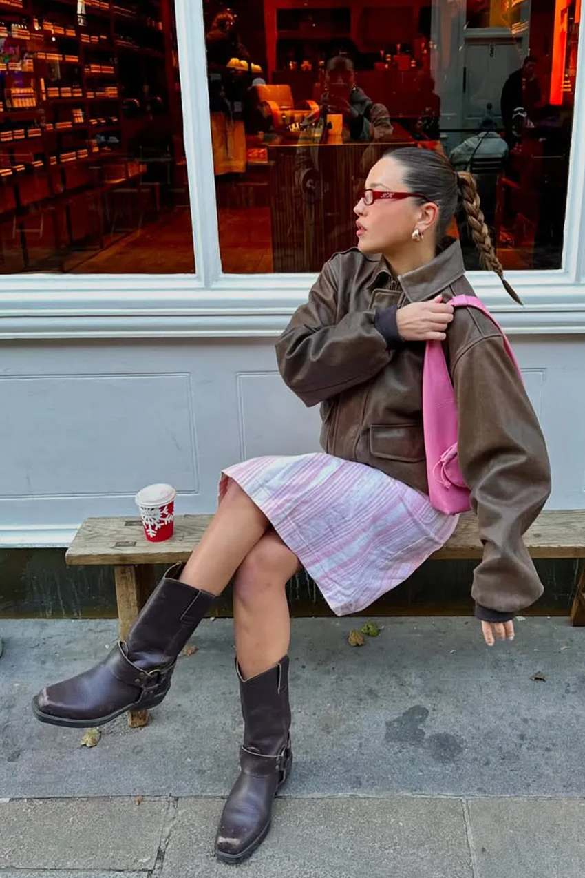 a woman wearing a brown leather jacket, pink shoulder bag, pink striped skirt, and brown boots
