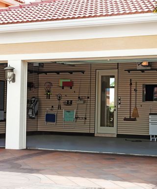 An house with a brick roof and beige walls with an open garage with light wooden wall panels with tennis rackets, fishing rods, and chairs on the wall, a white door, a concrete floor, and brick tiles in front of it