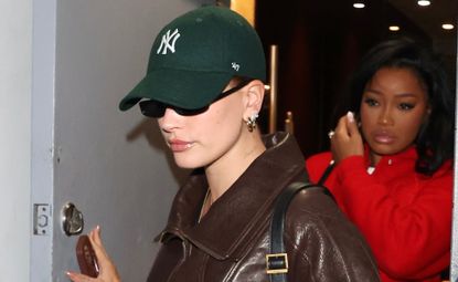 Hailey Bieber wears a chocolate brown bomber jacket with a glossy lip and baseball cap while out to dinner with a friend