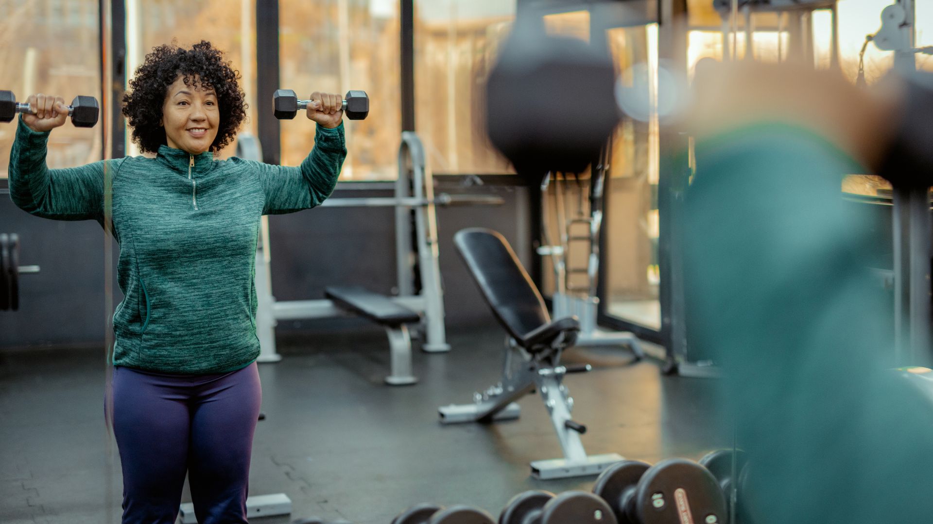 Woman lifting dumbbells in upper-body dumbbell workout at the gym, smiling and looking in the mirror