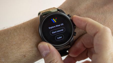 The Fossil Gen 6 smartwatch showing the initial screen "Explore Wear OS" with "Start tutorial" or "Later" buttons below. The wearer wears it on his left wrist and holds it lightly on both sides with thumb and index finger.