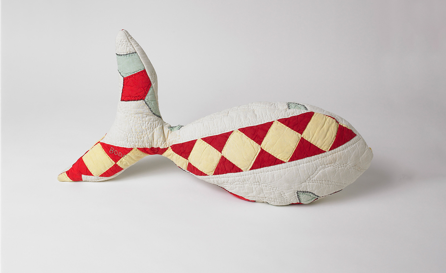 View of a multicoloured patchwork fish cushion with a diamond design by Emily Bode. The cushion is pictured against a light coloured background