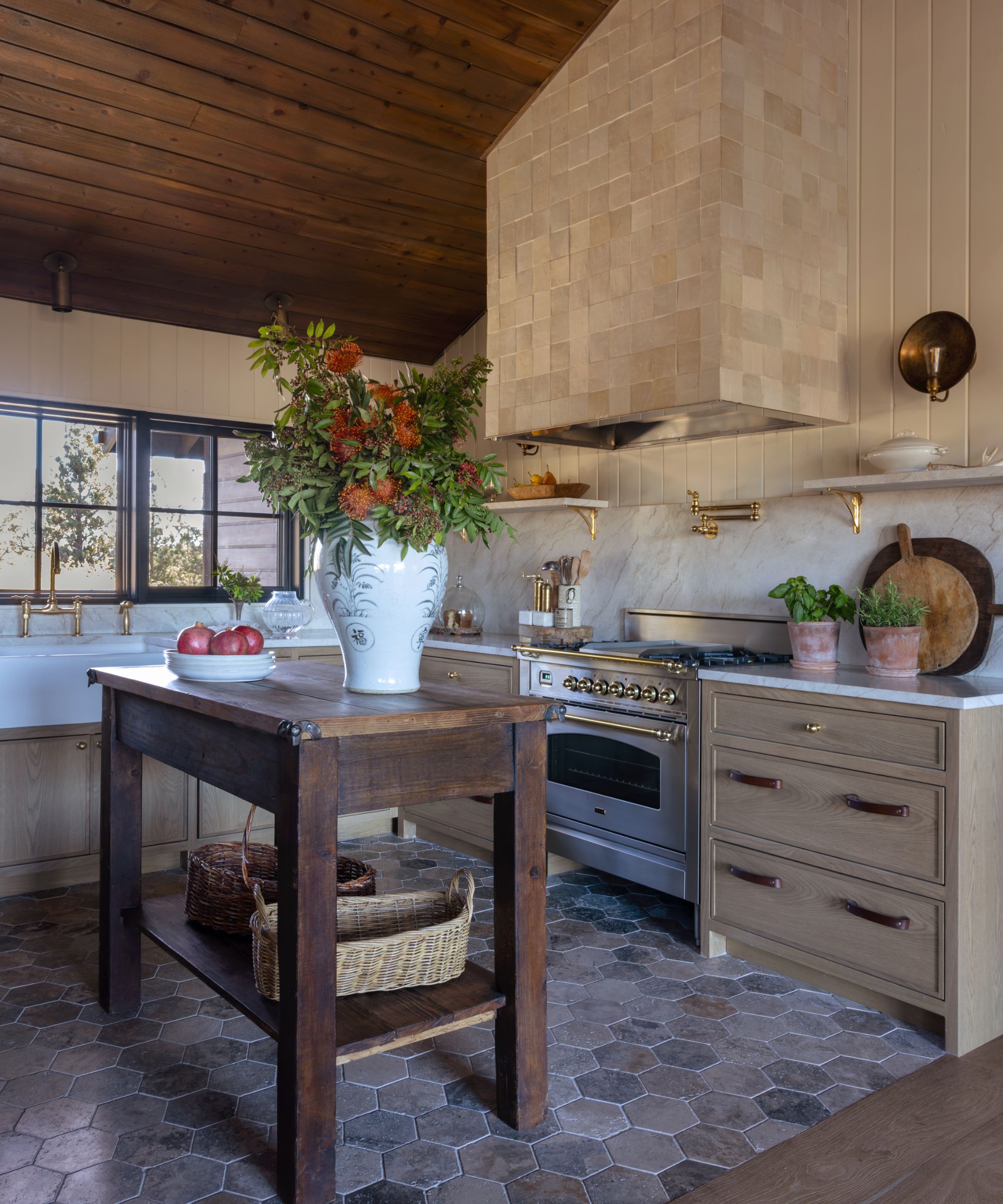A warm, rustic kitchen with wooden cabinets and a wood island, floral arrangement, open shelves, and hex tile flooring.