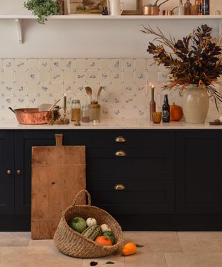 A kitchen with white and blue tiles, a countertop with candles, vases, and copper bowls on top, dark navy blue cupboards with gold handles, and a wooden chopping board with a basket of pumpkins next to it