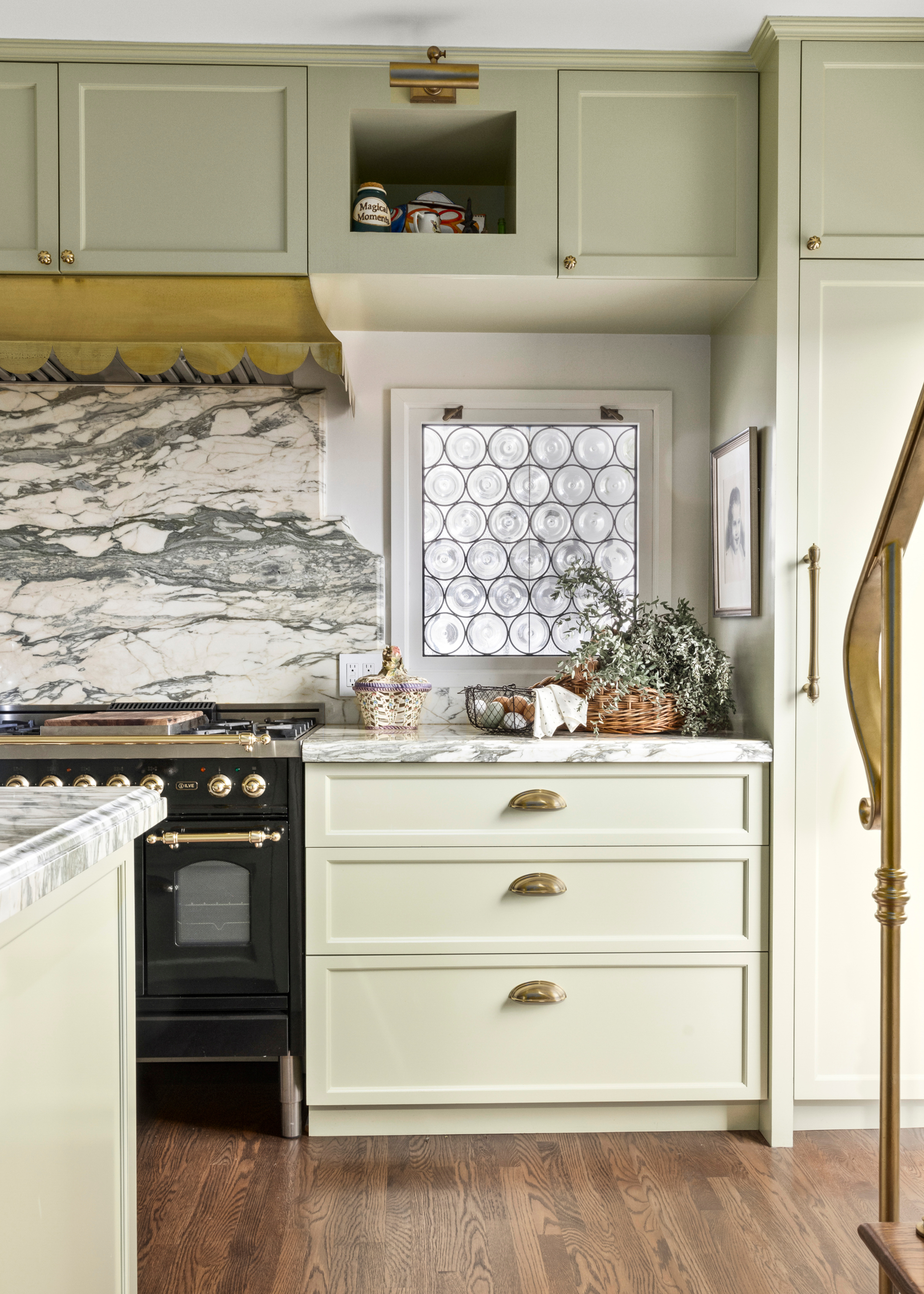 A kitchen with textured glass windows, light pastel green drawers, a stone backsplash and a scalloped exhaust