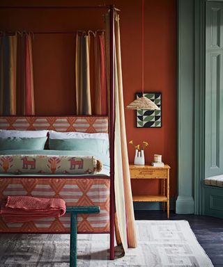 Four-poster bed with curtains, upholstered with orange fabric with pink geometric pattern and green bedding. Red painted wall behind bed, green painted wall on wall with windows