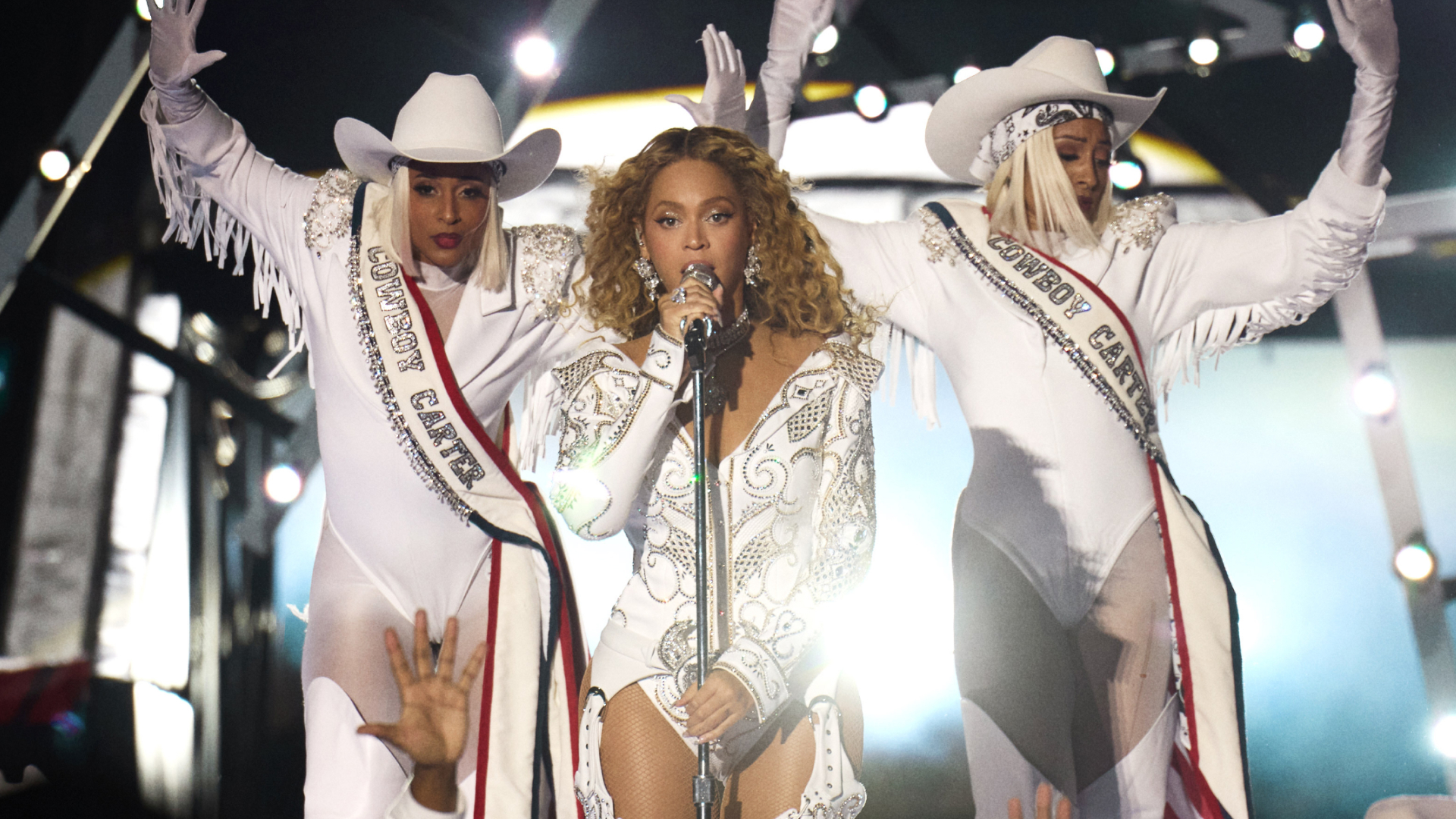 Beyoncé sings into a mic stand on stage with two backup dancers in white Western costumes, during Netflix's 'Beyoncé Bowl.'